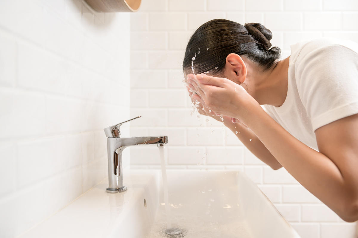 woman splashing water on her face over sink in a white tiled bathroom